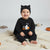 A baby wearing a black long-sleeve Halloween outfit with a matching black cat-ear hat, sitting on a cream blanket with plush toys in the background.