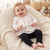 A baby sitting on a cream knit beanbag chair, wearing a white long-sleeve ruffle top with pink bow prints and black pants.