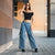 A woman stands on a paved walkway outdoors, wearing a fitted black off-the-shoulder crop top, wide-leg light blue denim cargo pants, and black shoes. She is posed between architectural columns with a blurred tan building in the background.