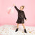 A young girl with blonde hair in a bun stands on a white fluffy rug, wearing a black sequined long-sleeve jacket and matching skirt with a black belt, black boots, and smiling as she reaches for a pink honeycomb paper balloon against a solid pink background.