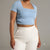 A model wearing a light blue short-sleeve crop top with a square neckline and white high-waisted pants, shown from the neck down against a solid gray studio background.