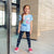 A young girl with glasses and a ponytail stands in front of a modern glass building, wearing a light blue graphic t-shirt with a cartoon character and the text 'Meow Time!', dark patterned leggings, and pink sneakers.