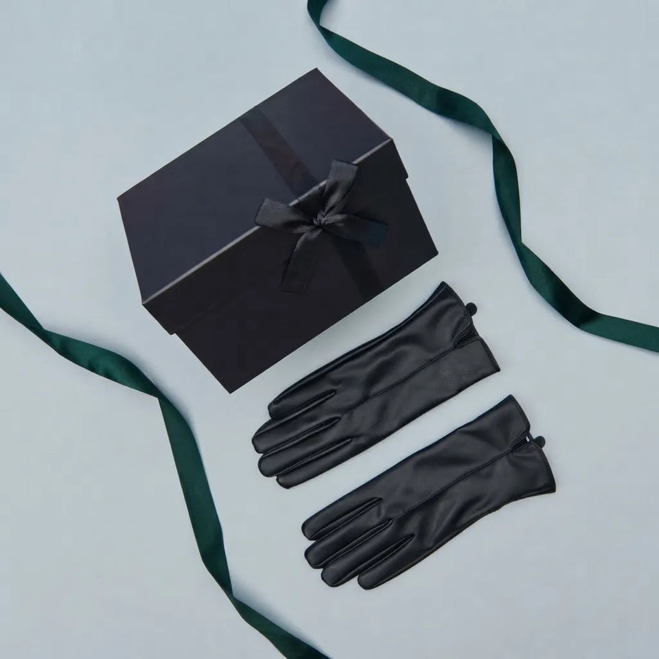 A pair of black leather gloves laid flat next to a black gift box tied with a dark green ribbon on a light gray background.