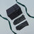 A pair of black leather gloves laid flat next to a black gift box tied with a dark green ribbon on a light gray background.