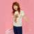 A young girl with red hair wearing a cream short-sleeve t-shirt with a brown poodle graphic and navy blue pants, posing against a solid pink background.