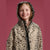 A young girl smiling while wearing a hooded rain poncho with a black and beige leopard print pattern, set against a solid dusty pink background.