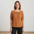 A woman modeling a rust-colored short-sleeve blouse with a gathered neckline and a small metallic accent, paired with black pants, against a neutral studio background.
