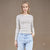 A woman modeling a fitted long-sleeve crewneck top with thin horizontal dark stripes on a white base, paired with light blue jeans, against a plain taupe studio background.