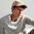A man wearing a beige baseball cap with a circular logo patch and a light gray ribbed knit sweater, photographed against a clean white background.