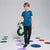 A young boy wearing a blue camouflage short-sleeve athletic t-shirt, black pants, and black sneakers, walking on a light gray studio floor decorated with colorful circular shapes.