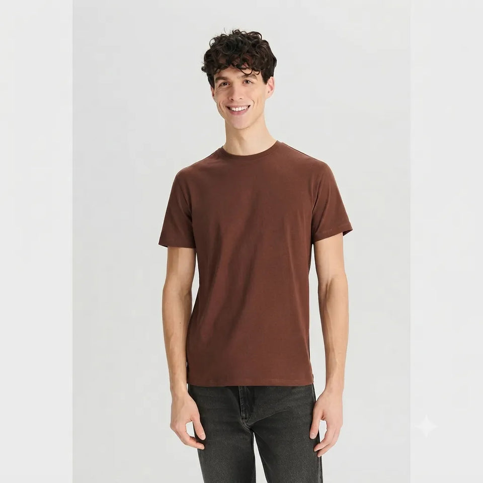 A man wearing a solid brown short-sleeve crewneck t-shirt and dark gray jeans, standing against a light gray studio background.