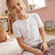 A young girl smiling while wearing a white short-sleeve t-shirt with delicate lace trim on the pocket and cuffs, paired with a floral pajama set, sitting on a pink carpet in a child's bedroom.