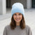 A smiling woman wearing a light blue fuzzy knit beanie hat and a gray knit sweater, photographed outdoors against a softly blurred neutral background.