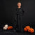 A child wearing a long black costume robe stands against a black studio background, with plush pumpkins placed on the floor around them.