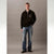 A male model stands against a neutral gray studio background wearing a black quarter-zip pullover sweater with a white embroidered Calvin Klein logo on the chest, paired with blue jeans and black shoes.