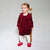 A toddler girl wearing a long-sleeve burgundy velvet dress with a large bow at the chest, white tights, and red shoes, standing on a wooden stool against a light gray studio background.