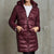 A woman wearing a shiny, quilted hooded puffer jacket in a deep burgundy/maroon color, standing against a textured gray concrete wall.