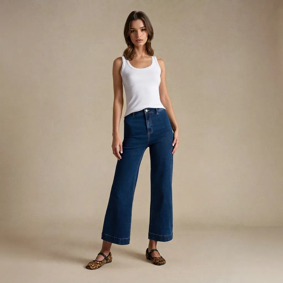 A woman stands against a plain beige studio backdrop wearing a white sleeveless tank top, dark blue wide-leg cropped jeans, and leopard-print flat shoes.