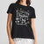 A woman wearing a black short-sleeve graphic T-shirt with 'Merry Christmas Days' and 'ALL I NEED IS HERE' printed in white and metallic script, surrounded by white snowflakes. The model is also wearing silver sequined bottoms against a light gray studio background.