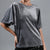 A woman wearing a gray short-sleeve velvet t-shirt with a relaxed, oversized fit and draped sleeves, paired with black sequined bottoms, against a solid gray background.