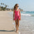 A young girl walking on a sandy beach next to the ocean, wearing a bright pink wrap-style one-piece swimsuit and sunglasses.