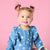 A smiling toddler girl with her hair in two pigtails wearing a blue long-sleeve floral romper, posed against a solid pink background.