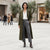A woman stands in a city plaza wearing a long olive green coat over a white blouse and gray trousers, with dark brown ankle boots. A Calvin Klein store is visible in the background.