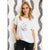 A woman wearing a white short-sleeve t-shirt with a black line art graphic of a crescent moon, stars, and a butterfly. She is posing in front of a white wall with gray abstract paper cutouts.