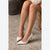 A close-up of a person's legs wearing light beige pointed-toe high heels with slim stiletto heels, seated on a wooden chair on a textured concrete floor.