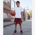A young boy stands on an outdoor basketball court wearing a white t-shirt, red and black patterned Nike shorts, a red headband, and red and black sneakers, holding a basketball.