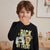 A young boy with brown hair wearing a black long-sleeve shirt with a 'Trick or Treat' graphic featuring a video game controller and a pizza slice, standing in a softly lit indoor room.