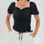 A woman wearing a black puff-sleeve top with a square neckline, ruffled trim, and front ruching, paired with off-white pants and a black belt, against a light gray studio background.