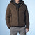 A man wearing a brown hooded puffer jacket over a black hoodie and gray sweatpants, shown from the neck down against a light blue studio background.