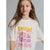 A young girl with long light brown hair wearing a white graphic t-shirt with colorful retro-style text that reads 'Spread good energy like super sun glow happiness' in yellow, orange, pink, and purple, set against a plain gray studio background.