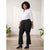 A woman stands indoors modeling a white button-up shirt and black flared pants with a black belt, paired with black ankle boots. The setting has a light neutral wall, concrete floor, and potted plants in the background.