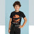 A young boy with curly brown hair wearing a black graphic t-shirt with an orange and black 'Scary Rex' dinosaur design, paired with dark jeans and a checkered belt, posing against a light blue background.