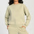A person wearing a light sage green crewneck sweatshirt and matching sweatpants, shown from the neck down against a plain light gray studio background.
