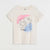 An off-white short-sleeve t-shirt with a graphic print of a woman's face and cat in black line art, set against pink and blue watercolor shapes, with the word 'dream' written below.