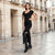 A woman posing on a wet cobblestone street in front of a classic beige stone building, wearing a black short-sleeve ribbed scoop-neck maxi dress with a thigh-high slit and black over-the-knee boots.