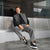 A man sits on a modern bench wearing a gray sleeveless zip-up vest over a black shirt, gray wide-leg jeans, and black sneakers with white laces.