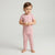 A toddler standing barefoot against a light gray studio background, wearing a matching pink short-sleeve top and leggings set with small white polka dots.