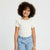 A young girl with curly brown hair wearing a cream ribbed short-sleeve top with ruffle trim and light blue polka dot pants, standing against a plain white background.
