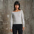 A woman modeling a light gray off-the-shoulder long-sleeve knit sweater with black pants, standing against a textured concrete wall.