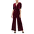 A woman modeling a deep burgundy velvet jumpsuit with short sleeves, a wrap-style V-neckline, and a twisted waist detail, paired with black pointed shoes against a plain white background.