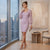 A woman models a light purple ribbed knit mini dress with long balloon sleeves in a modern room with a city view, holding a small silver clutch.