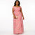 A woman modeling a pink off-the-shoulder maxi dress with a floral watercolor print, holding a small beige clutch against a plain white studio background.