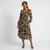 A woman modeling a long-sleeve midi dress with a dark floral print, featuring a V-neckline, tie waist, and flowing skirt, set against a plain light gray studio background.