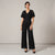 A woman modeling a black lace short-sleeve jumpsuit with wide legs, standing against a neutral light gray studio background.