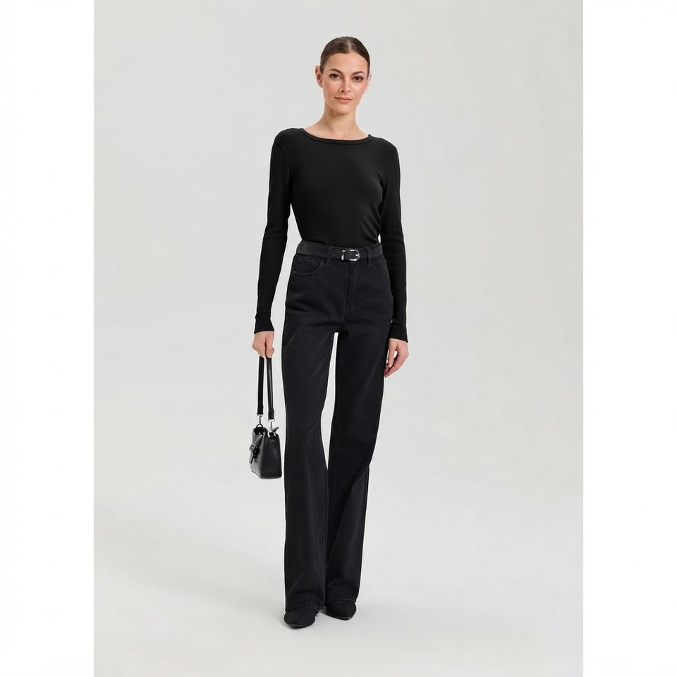 A woman modeling a black long-sleeve boat-neck top and black high-waisted wide-leg jeans, styled with a black belt and holding a small black handbag, against a light gray studio background.