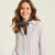 A woman with wavy brown hair smiling, wearing a white long-sleeve button-up shirt with thin black vertical pinstripes and a black-and-white patterned neck scarf, set against a plain light beige studio background.